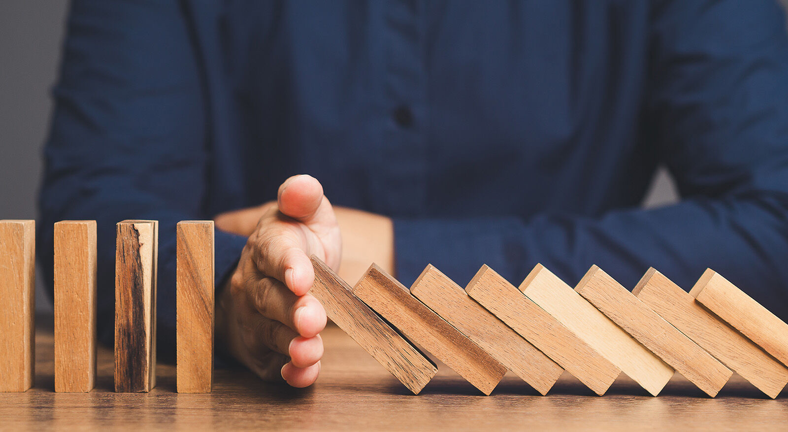 Hand stoppt fallende Dominosteine auf einem Holztisch vor einer Person in blauem Hemd