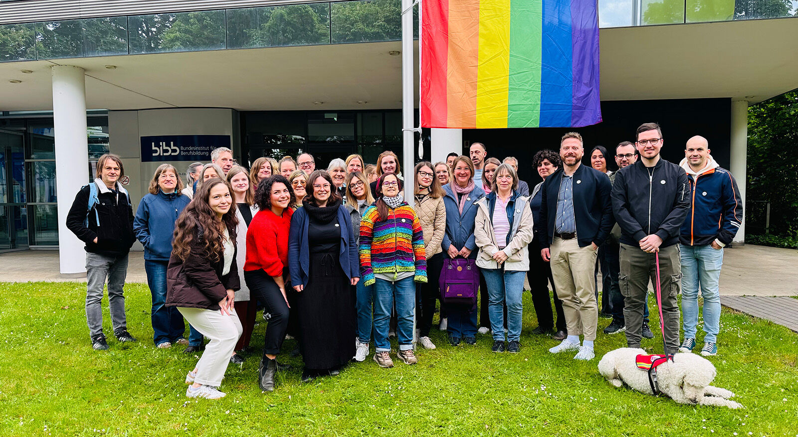 Gruppe von Menschen vor einem Geb&auml;ude mit gro&szlig;er Regenbogenflagge und kleinem wei&szlig;en Hund auf Grasfl&auml;che