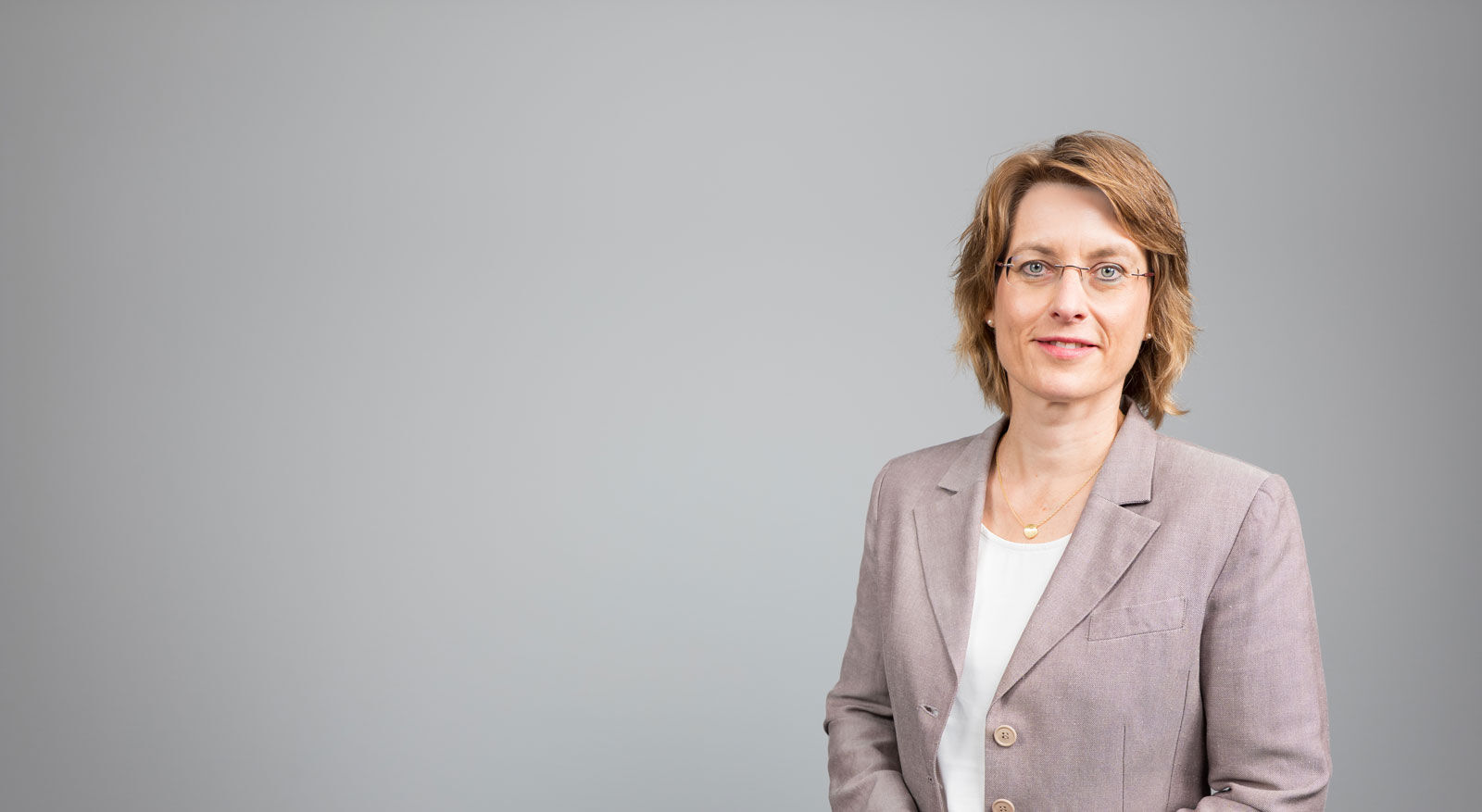 Frau mit kurzem Haar und Brille in hellem Blazer vor grauem Hintergrund