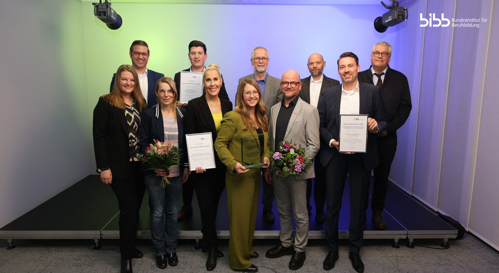 Eine Gruppe von elf Personen in formeller Kleidung auf einer B&uuml;hne, einige halten Urkunden und Blumenstr&auml;u&szlig;e