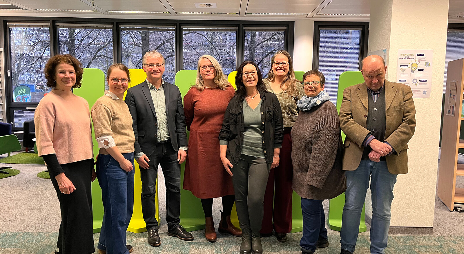 Eine Gruppe von acht Personen steht in einem modernen B&uuml;ro mit gro&szlig;en Fenstern und gr&uuml;nen Sitzm&ouml;beln im Hintergrund