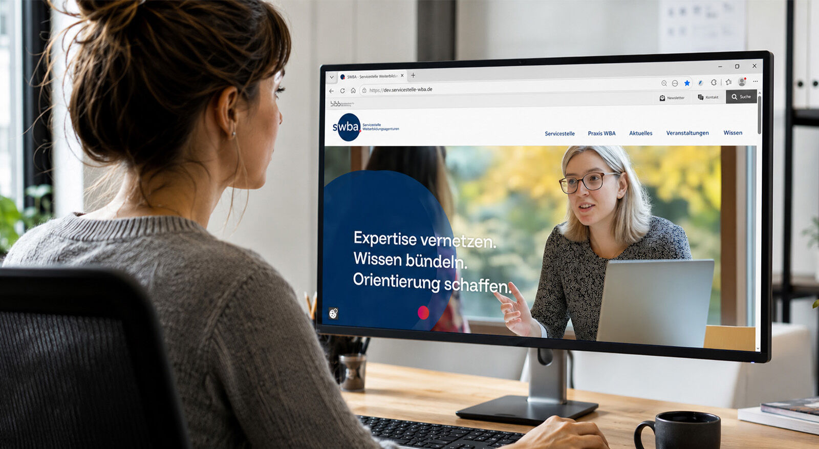 Frau sitzt am Schreibtisch vor einem Monitor, auf dem eine Webseite mit zwei Frauen und dem Text 'Expertise vernetzen Wissen b&uuml;ndeln Orientierung schaffen' zu sehen ist