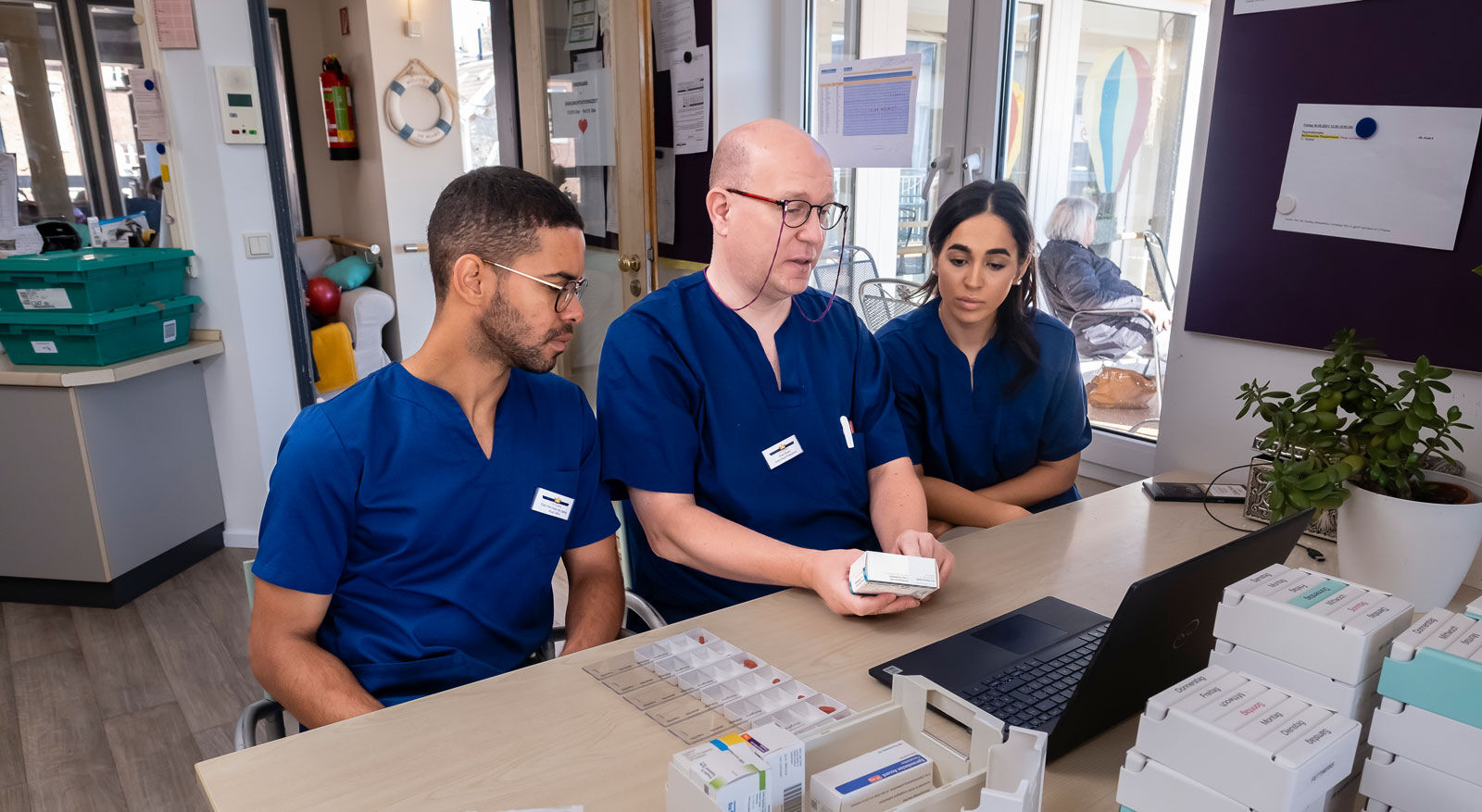 Drei Pflegekr&auml;fte in blauer Arbeitskleidung sitzen an einem Tisch mit Medikamentenpackungen und einem Laptop.