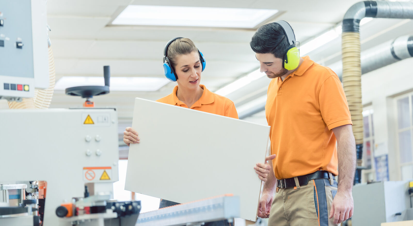 Woman explaining something to a man inside a workshop, both wearing hearing protection.