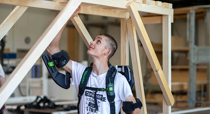 Ein Auszubildender trägt ein Exoskelett und arbeitet an einem Holzbauelement